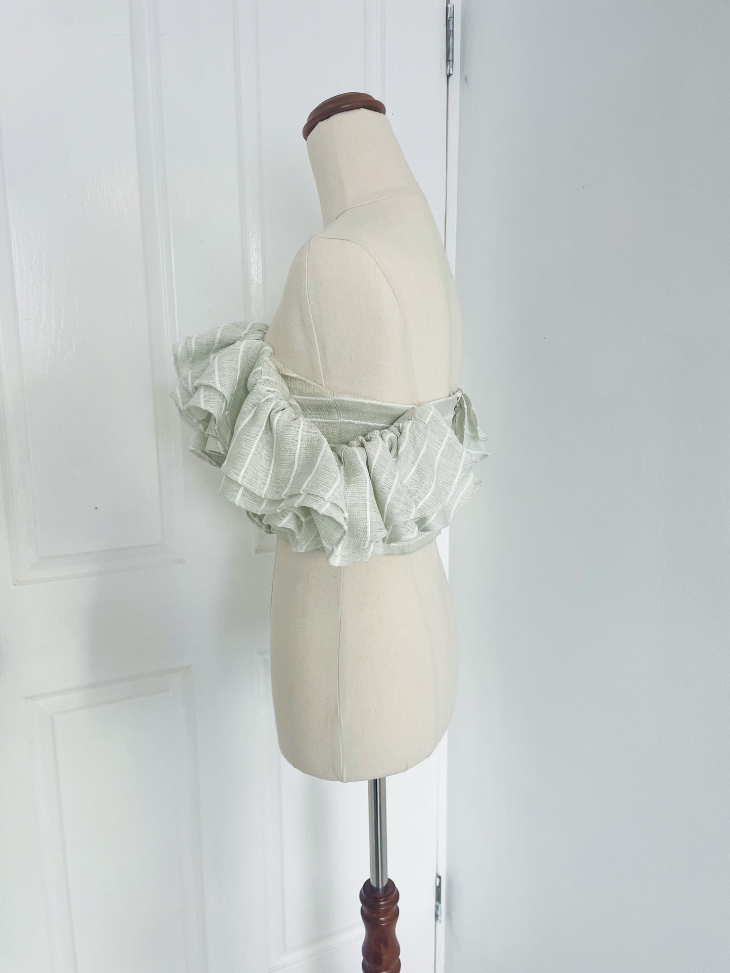 side view of a white mannequin displays a mint green, pinstripe , off the shoulder crop top with layered ruffles. designed by Acler.