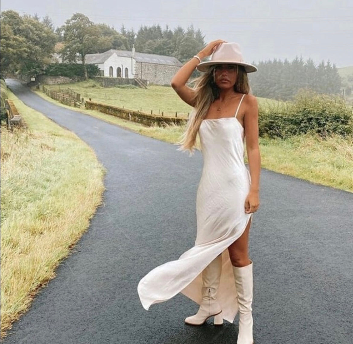 A country town, a woman is wearing a white satin gown with a thigh high split and below the knee white faux leather chunky heel boots she has her hand on her brim hat and wearing a bangle